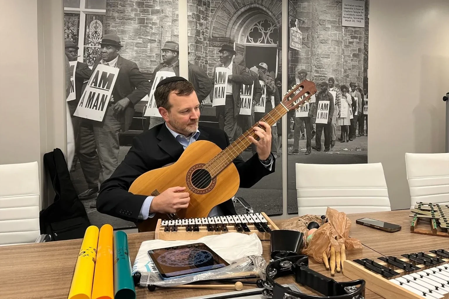 Jason Caplan playing guitar at a Universal Language Room table with simple instruments arranged for participation.