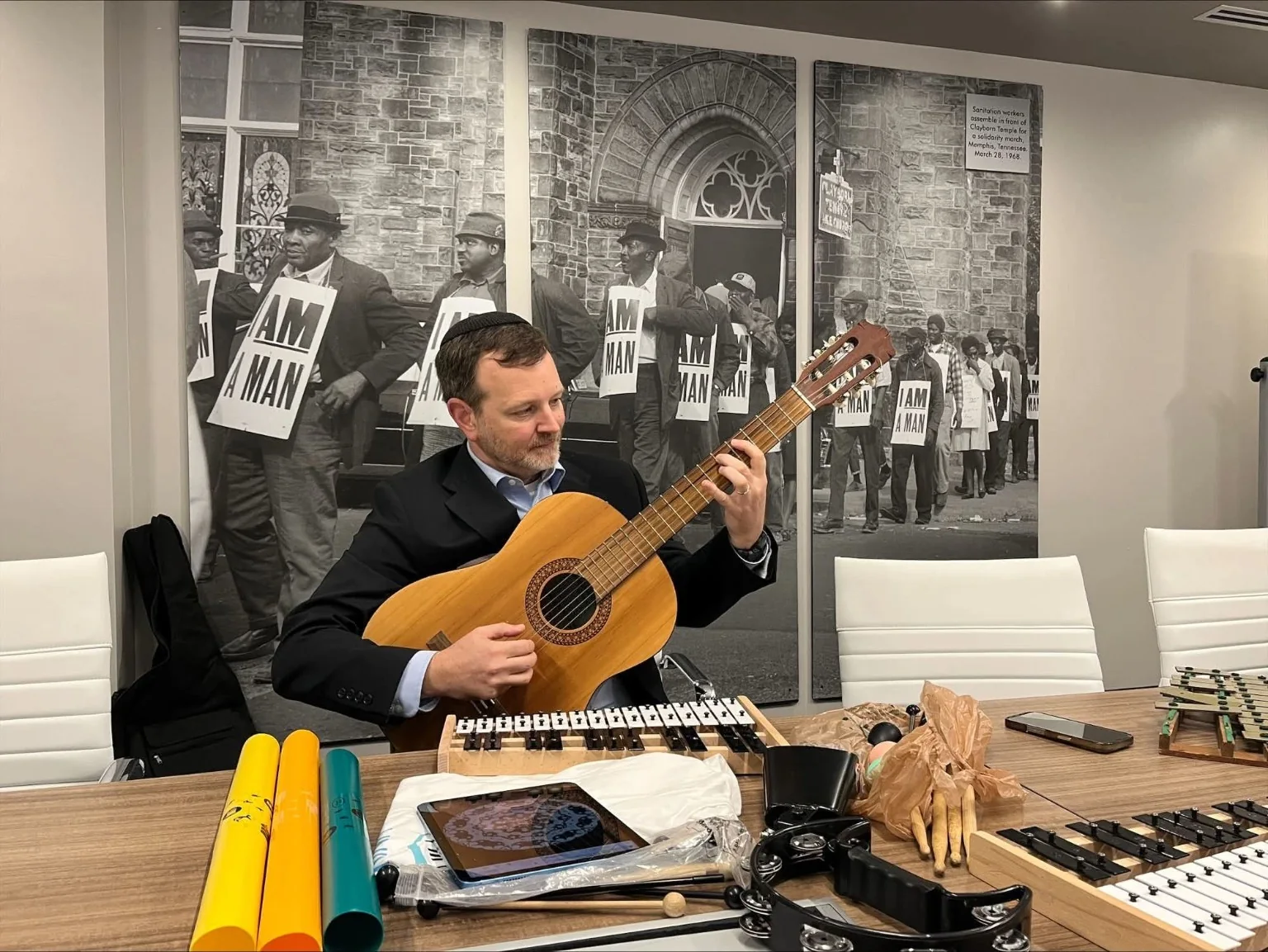 A facilitator playing guitar at a Universal Language Room table with simple instruments arranged for participation.